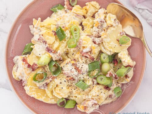 A square photo with a pink plate with salad of potatoes, a creamy dressing, Cheddar and bacon. A white background.