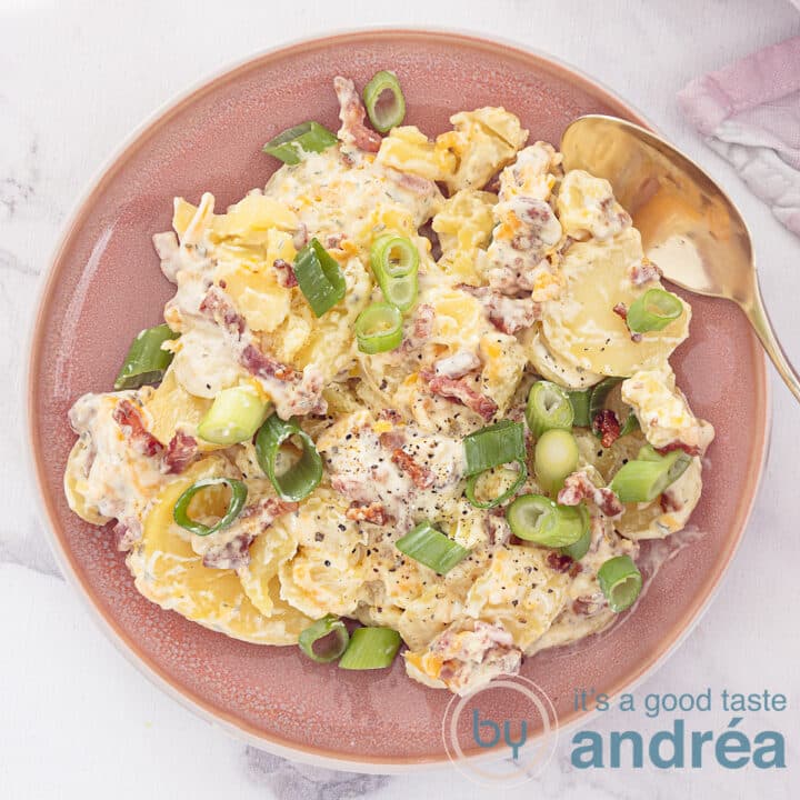 A square photo with a pink plate with salad of potatoes, a creamy dressing, Cheddar and bacon. A white background.