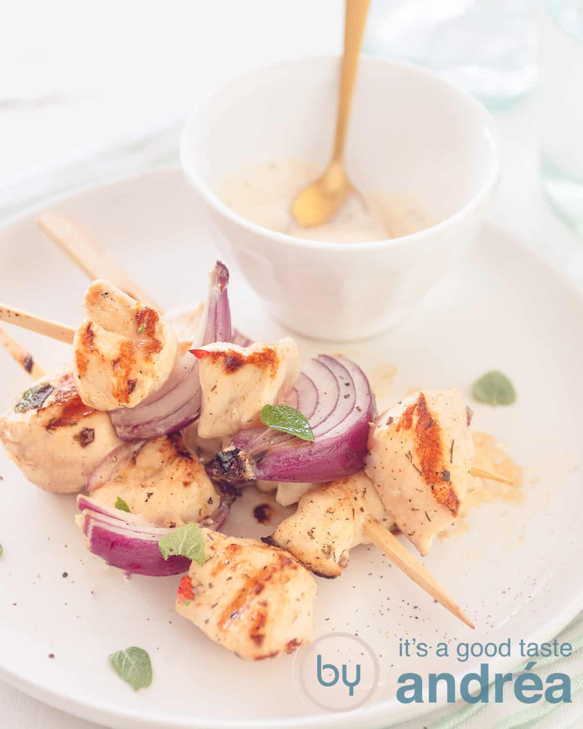 Een hoogtefoto met een wit bord op een witte ondergrond met een kommetje tzatziki. Op het bord drie kipspiesjes met kip afgewisseld met rode ui.