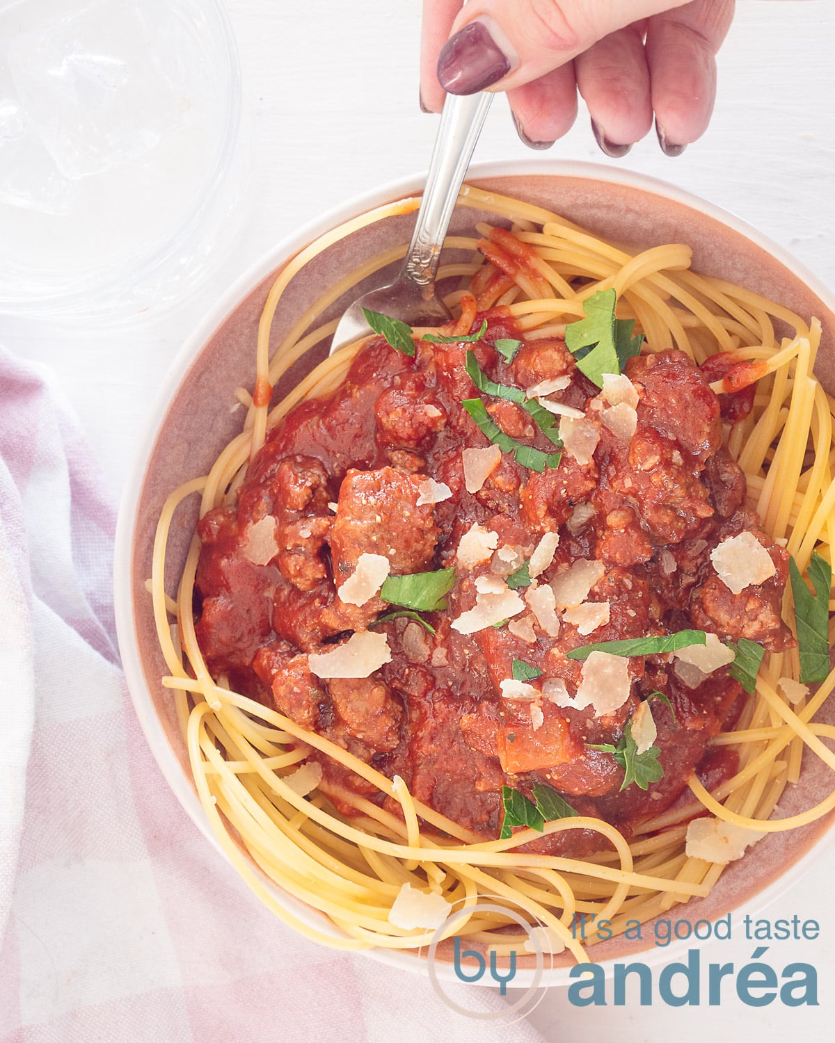 Een foto van bovenaf van een schaal met spaghetti met daarop zelfgemaakte bolognaisesaus met Parmezaanse kaas en peterselie. Een hand houdt een lepel vast. Een witte ondergrond, links onderin een wit roze theedoek.