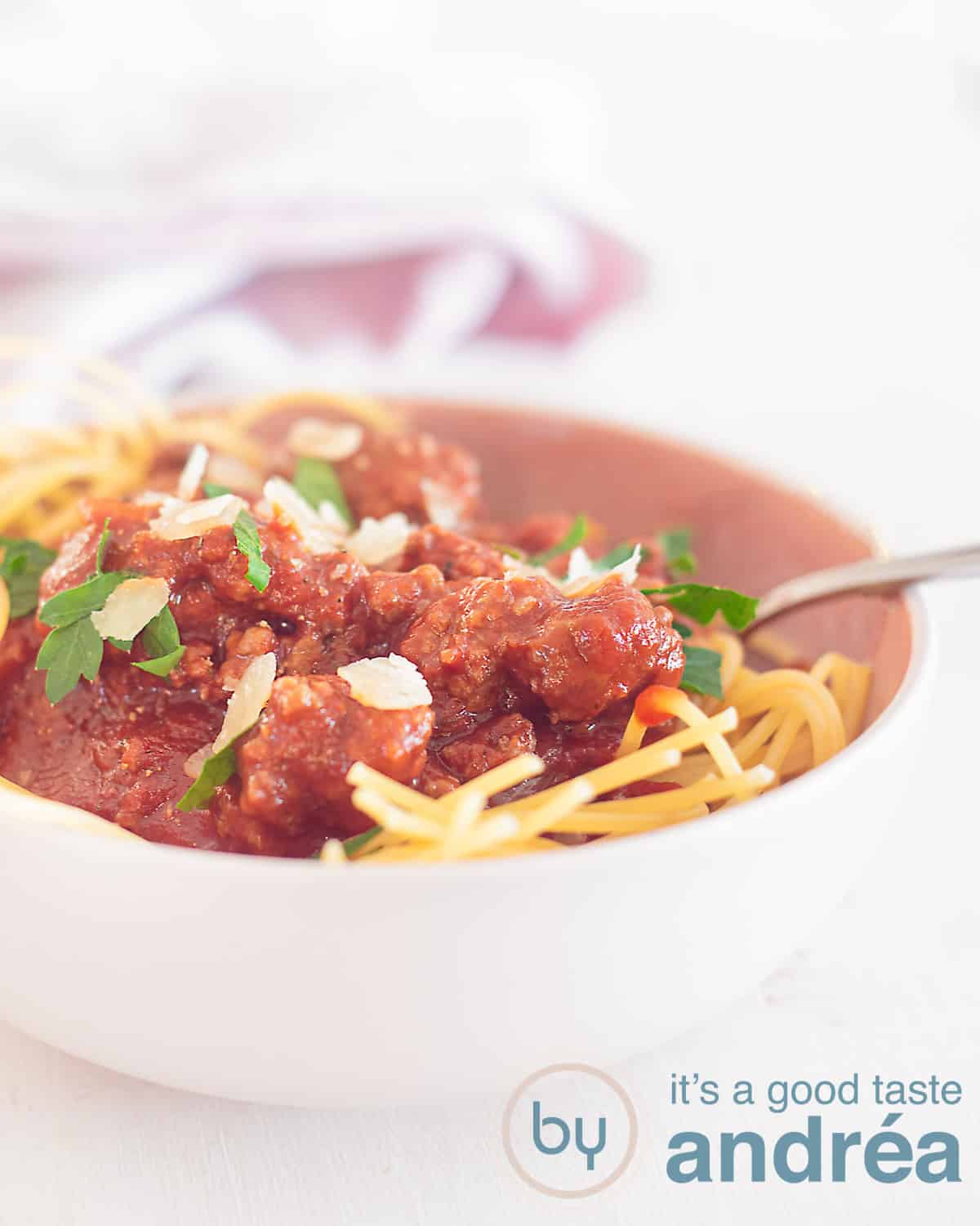 Een hoogtefoto met een roze schaal gevuld met spaghetti getopt met klassieke bolognesesaus. Een witte ondergrond. In de achtergrond een roze theedoek