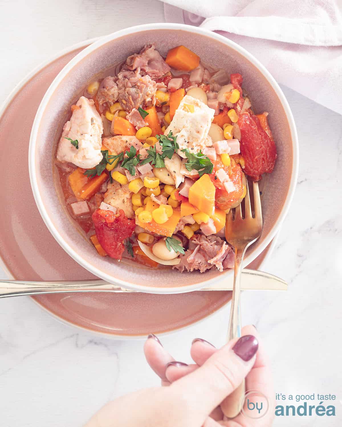 Een foto van bovenaf van een kom gevuld met kleurrijke Brunswick stew op een roze bord. Een witte ondergrond. Een hand houdt een vork vast die wat stoofschotel wil pakken.