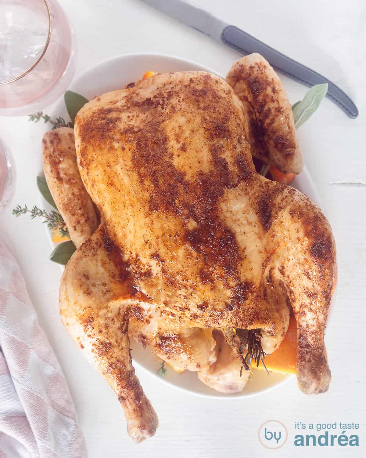 Geroosterde kip met Honing en Sinaasappel uit de oven op een wit bord. Van bovenaf gefotografeerd. Rechts bovenin een mes, links bovenin een roze glas.