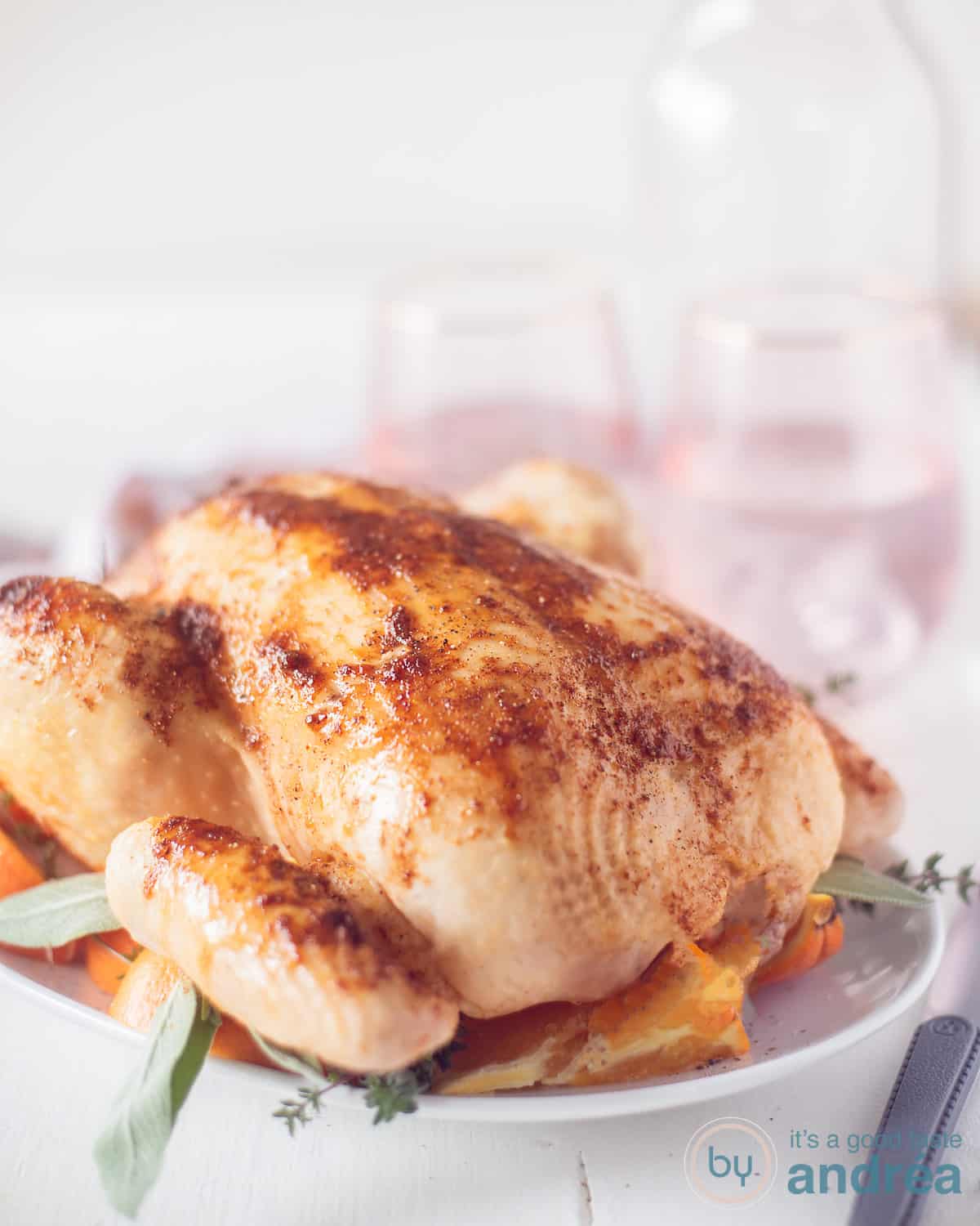 Geroosterde kip uit de oven op een bedje van sinaasappels en verse kruiden op een wit bord. In de witte achtergrond zie je twee roze glazen.