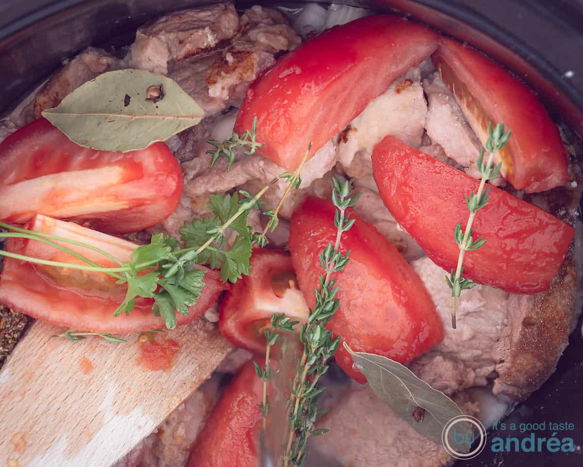 Een slowcooker met hamlappen, verse kruiden, tomaten en laurier
