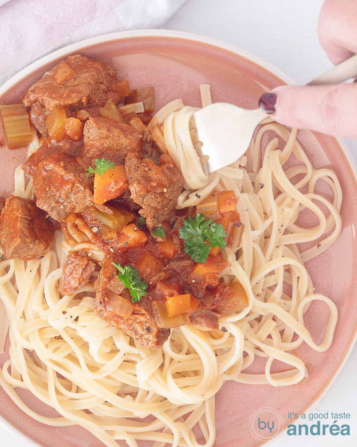 Een foto van bovenaf van een roze bord met tagliatelle met Italiaans stoofvlees uit de slowcooker. Een vork steekt in een stukje vlees.