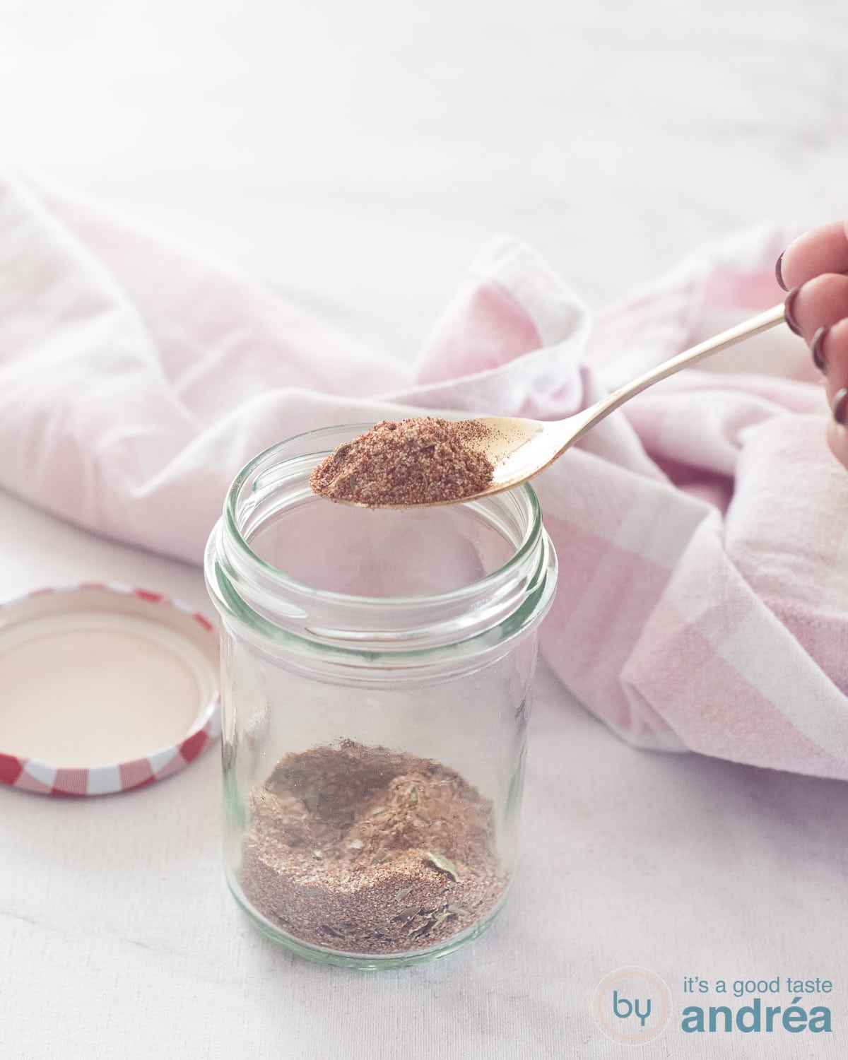 Een glazen pot met kipkruiden, een gouden lepel schept een lepel zelfgemaakte mix eruit. Een roze witte theedoek achterin.