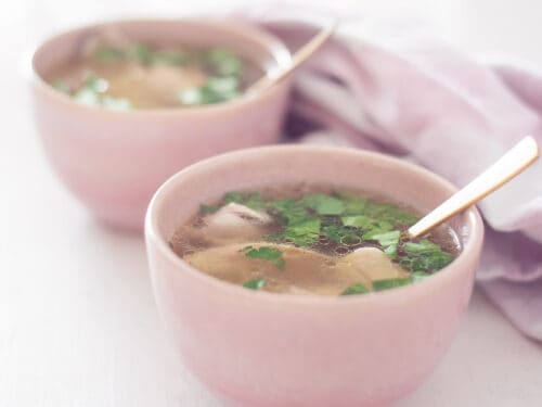 A square photo with two pink bowls filled with homemade fresh chicken soup. A white background and on the right a pink and white tea towel