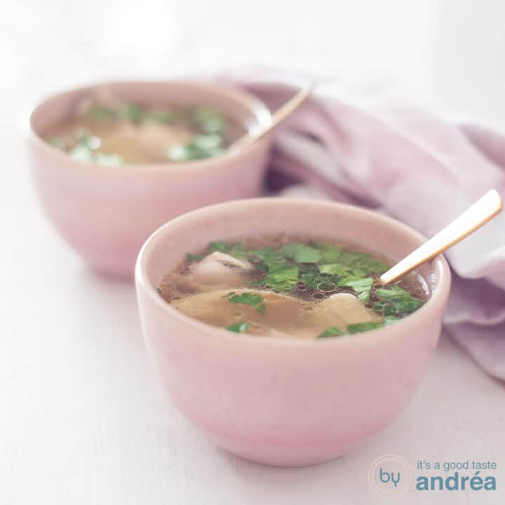 A square photo with two pink bowls filled with homemade fresh chicken soup. A white background and on the right a pink and white tea towel