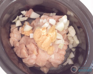 Een slowcooker met kipblokjes, kip, knoflook en ui.