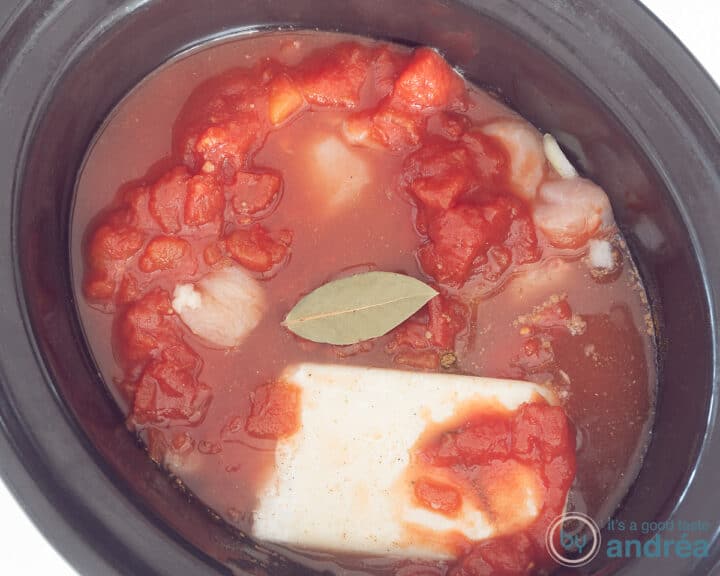 Een slowcooker met kip, kruiden, kokos en tomatenblokjes. Een laurierblad drijft in het midden.