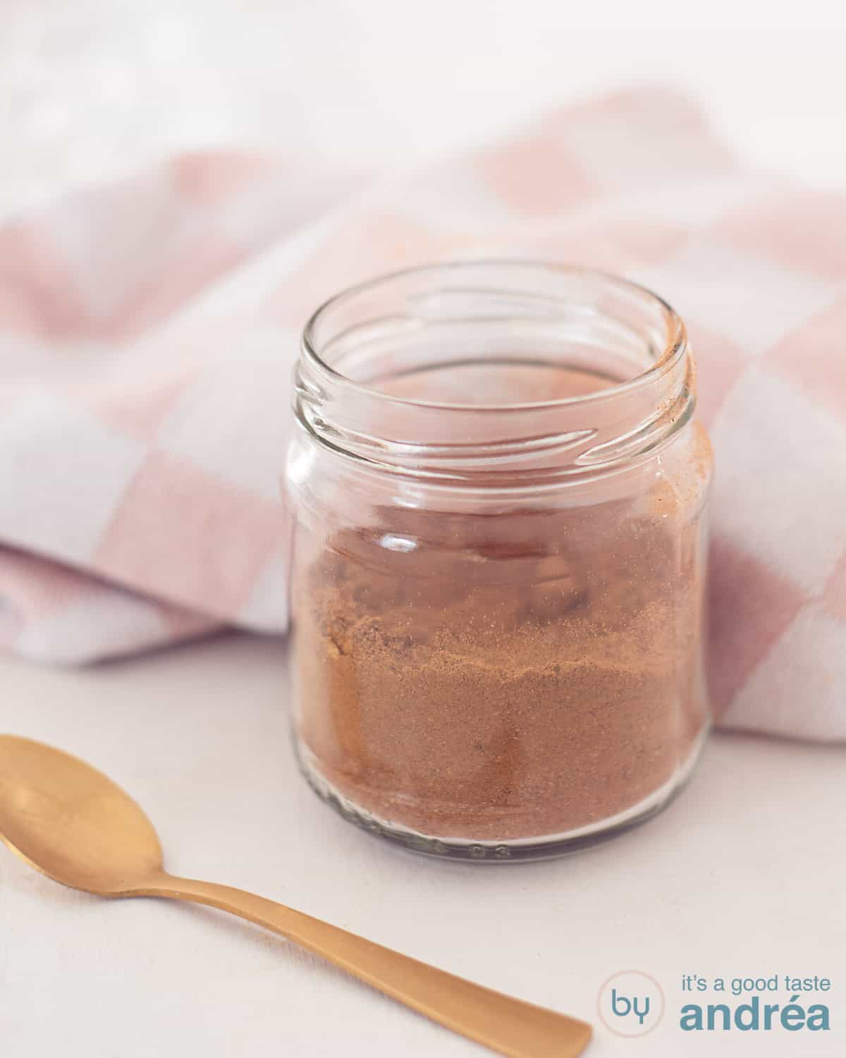 Een glazen potje gevuld met koekkruiden mix op een witte ondergrond. Een gouden lepel ligt ervoor. Een roze witte theedoek ligt erachter.