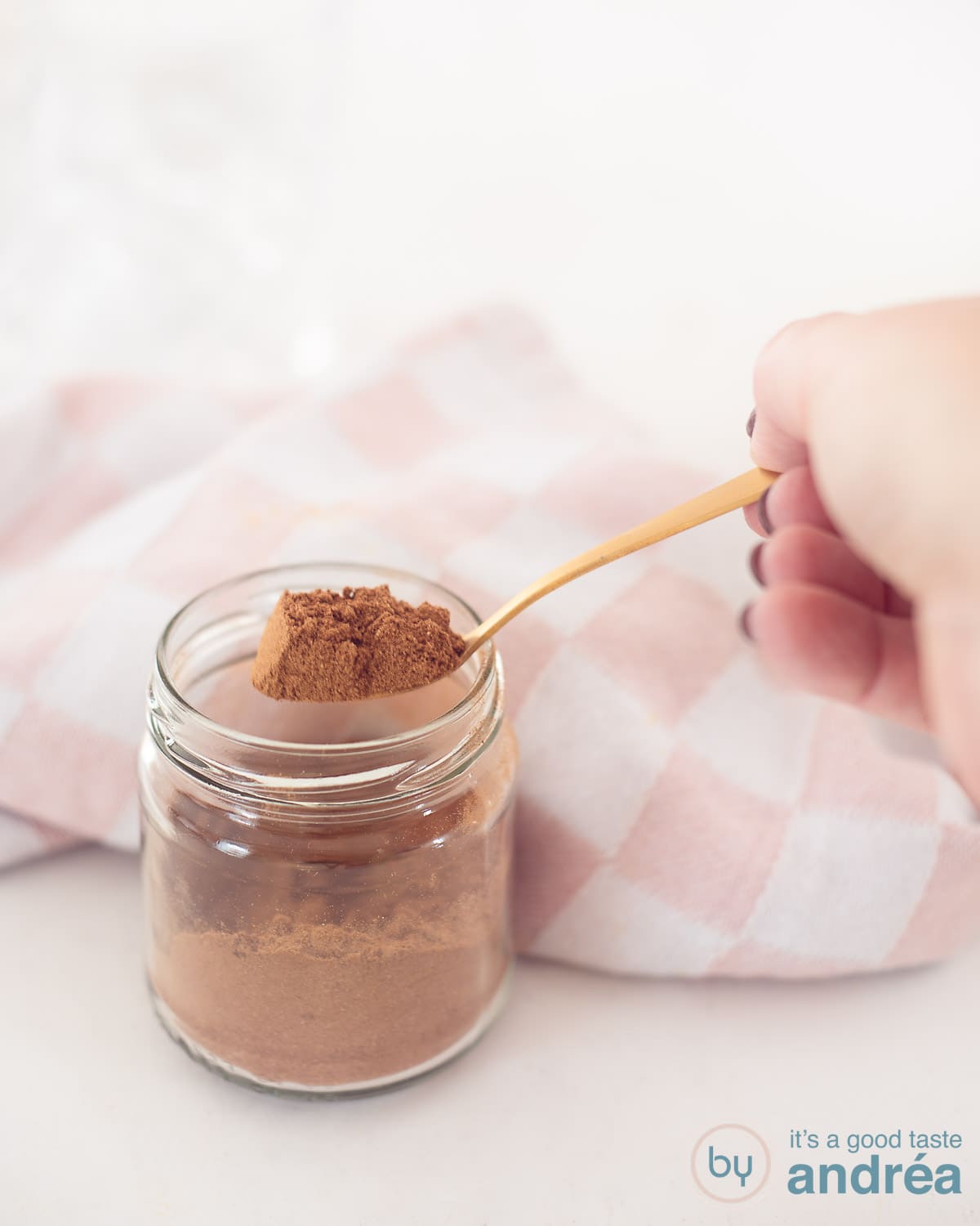 Een glazen potje met speculaas kruiden met een gouden lepel die wat mix op de lepel heeft. Een witte ondergrond. In de achtergrond een roze witte theedoek