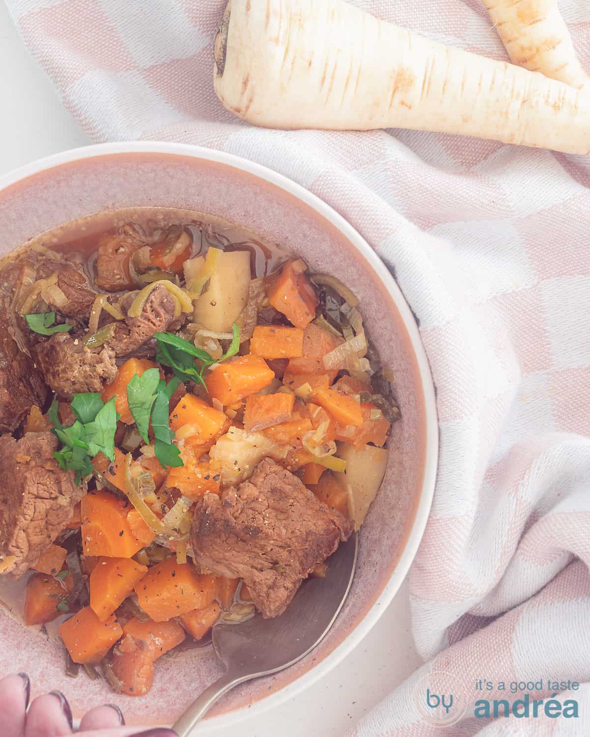 Een foto van bovenaf met linksonderin een roze kom met gestoofd rundvlees met wortel, zoete aardappel en pastinaak in een bier bouillon saus.