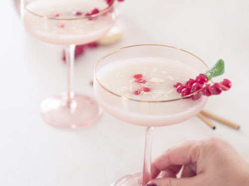 Een vierkante foto met twee roze champagneglazen gevuld met scroppino, granaatappelpitjes, rode bessen en munt. Een hand pakt het voorste glas.