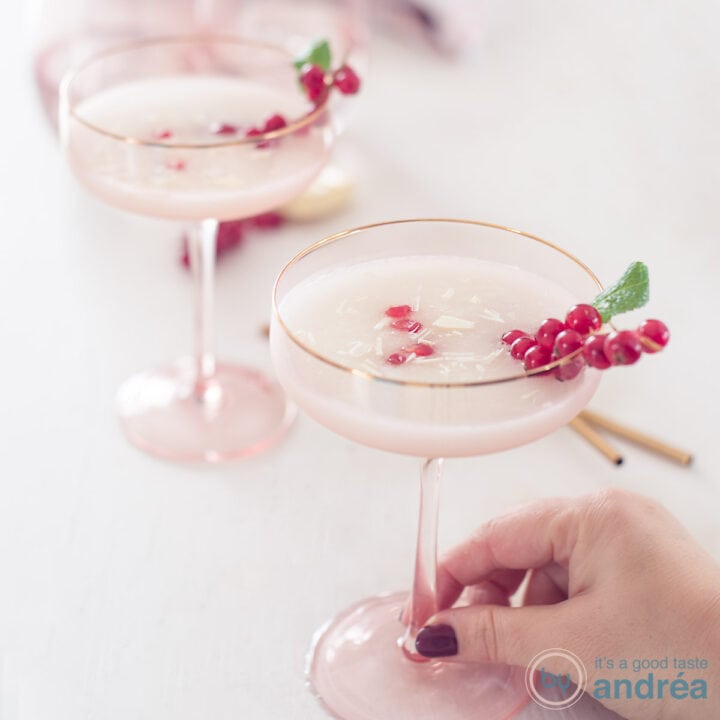 Een vierkante foto met twee roze champagneglazen gevuld met scroppino, granaatappelpitjes, rode bessen en munt. Een hand pakt het voorste glas.