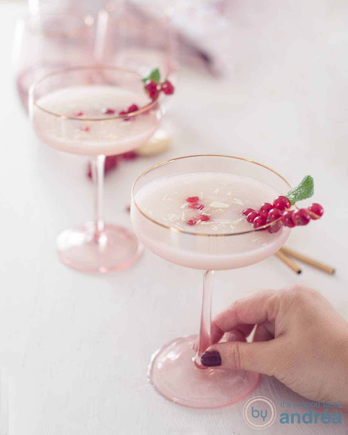 Een hoogte foto van twee roze champagneglazen gevuld met alcoholvrije spoom sgroppino. Gegarneerd met granaatappelpitjes, rode bessen en munt. Op een witte ondergrond.