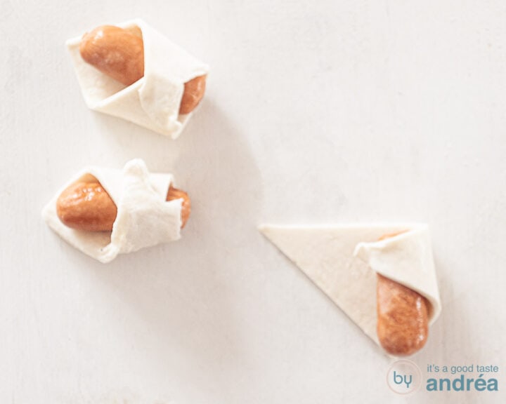 Twee cocktailworstjes gerold in bladerdeeg op een witte ondergrond. Een derde driehoek bladerdeeg bevat een cocktailworst waaromheen een punt bladerdeeg is gevouwen.