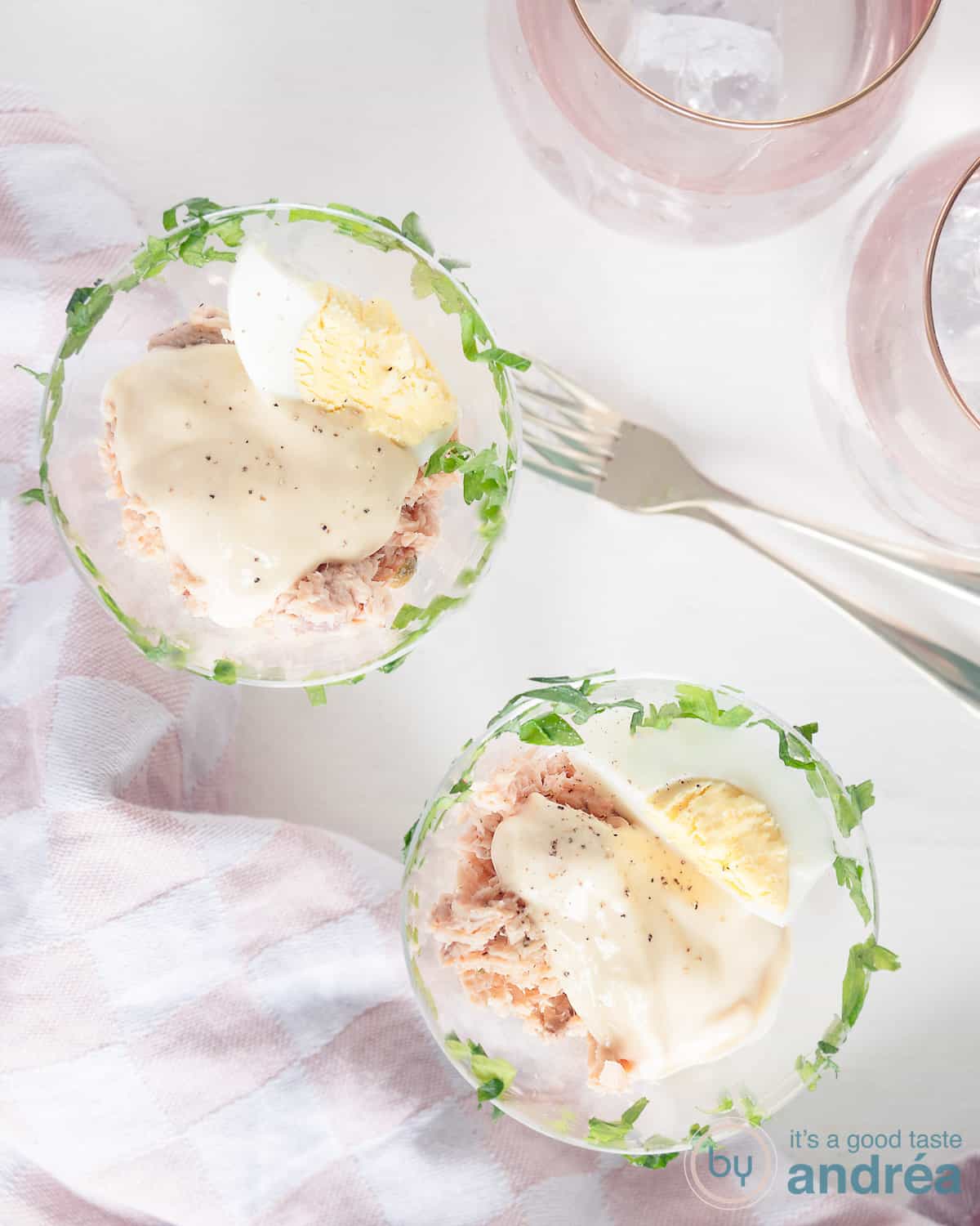 A photo from above with two glasses filled with whiskey salmon cocktail with egg. Two pink glasses at the top right. A pink and white tea towel at the bottom left