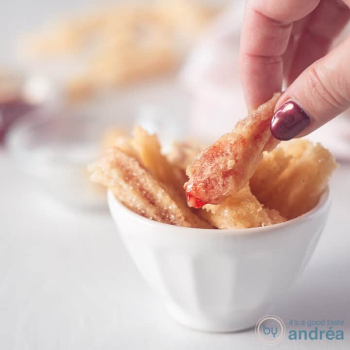 Een vierkante foto met een witte kom gevuld met aardbeienchurros. Een hand pakte een churro vast.