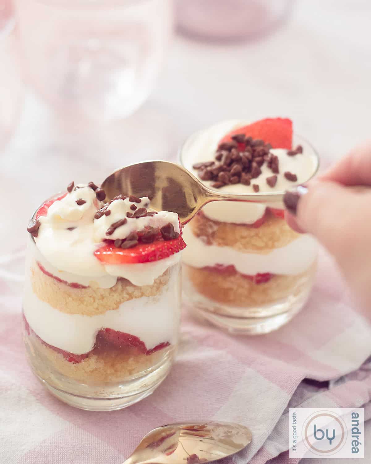 Twee glaasjes gevuld met aardbeien mascarpone room en cake trifles. Een gouden lepel neemt een hap.