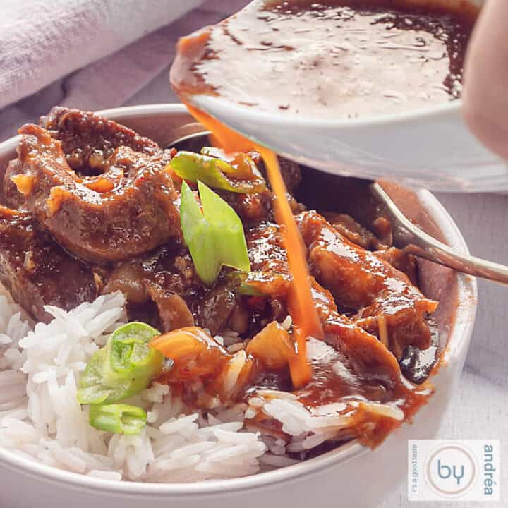 Een vierkante foto met een kom gevuld met babi pangang, rijst en een garnering van lente ui. Een witte om met saus giet extra saus over het vlees