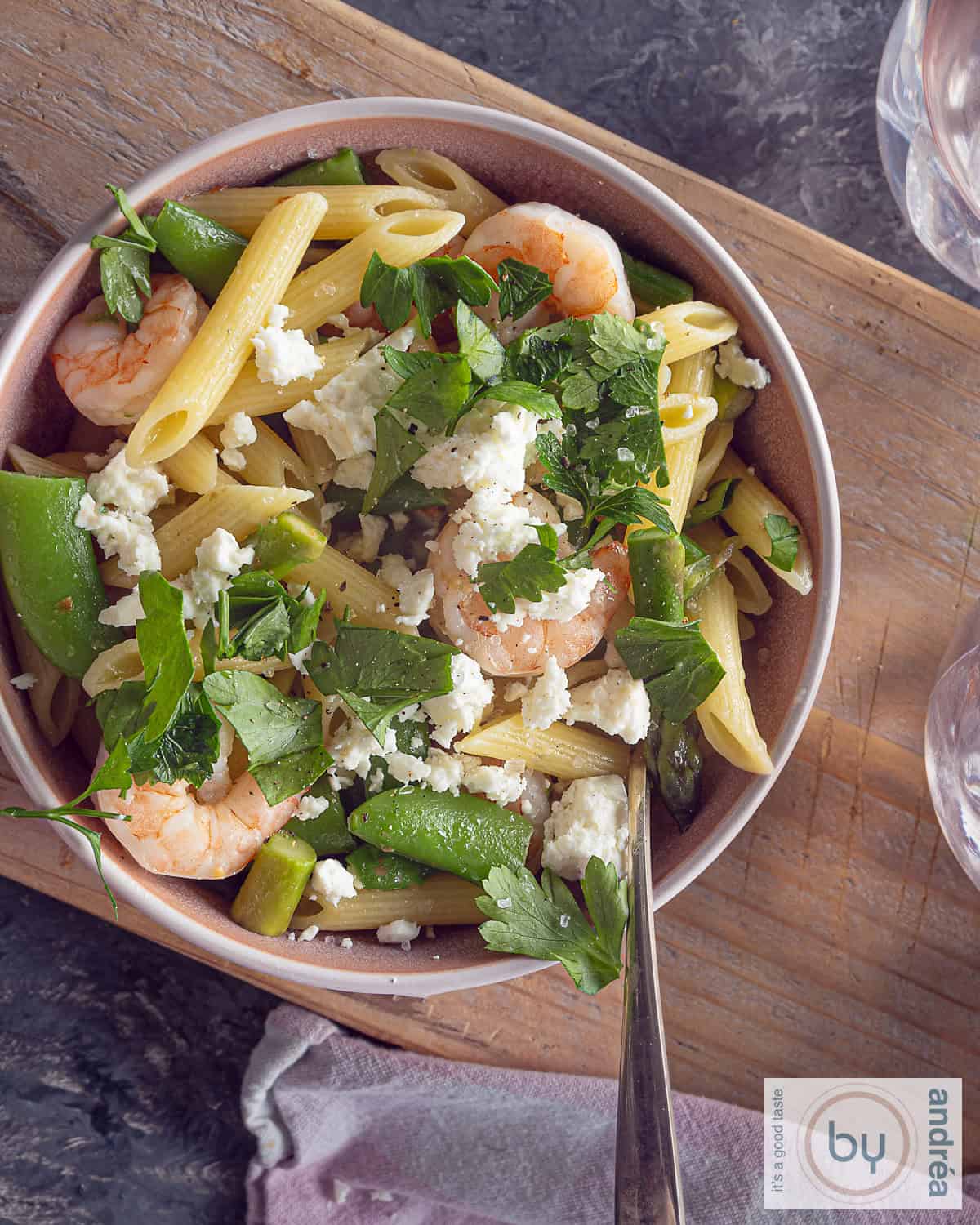 Een foto van bovenaf van een roze schaal met pasta, garnalen, lentegroenten, feta en peterselie. Alles op een houten plank.