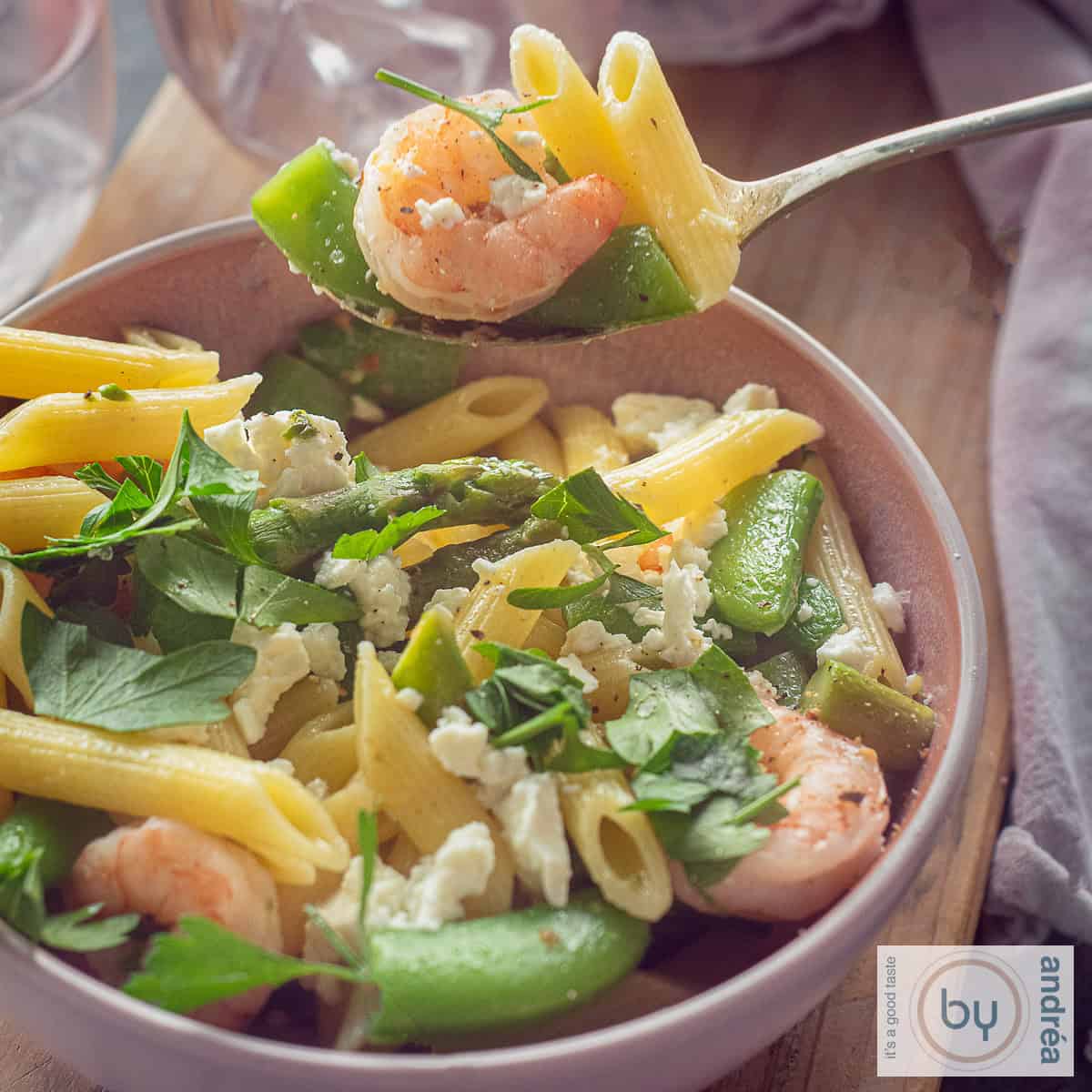 Een vierkante foto van een roze schaal met een mix van garnalen, penne en lente groenten. Een lepel schept een hap eruit.