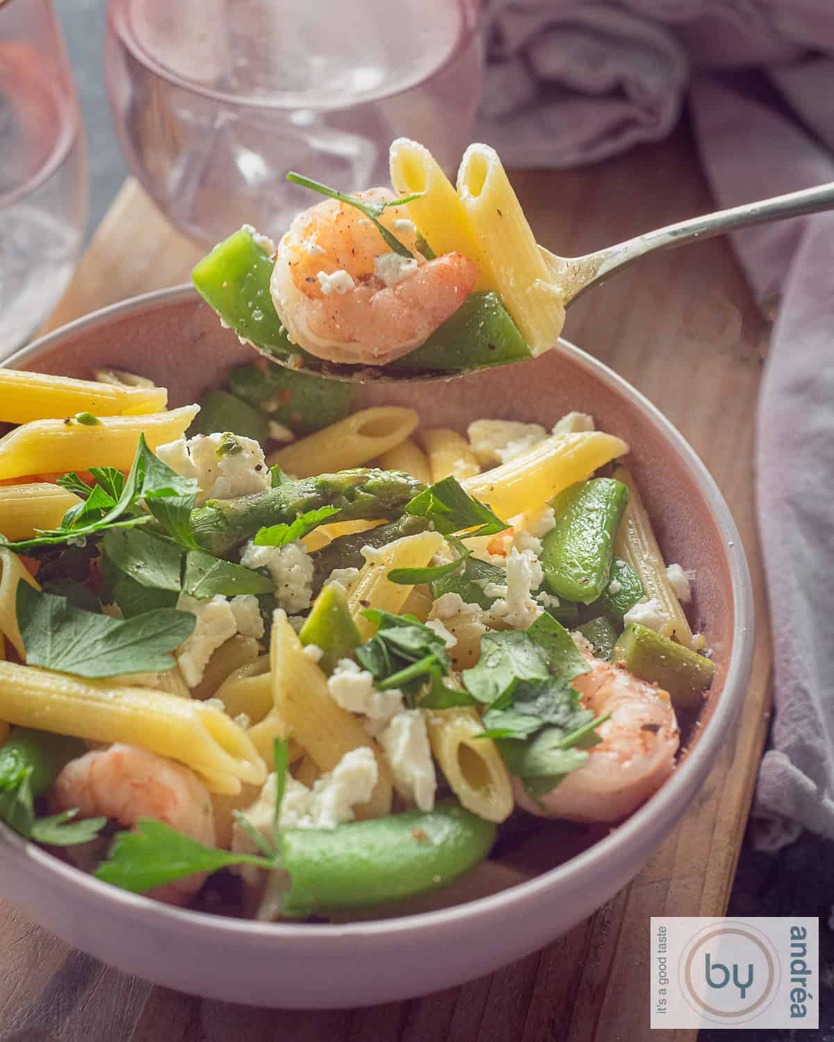 Een hoogtefoto met een roze schaal op een houten plank gevuld met pasta, garnalen, sugar snaps en asperges. Een lepel schept een hap eruit.