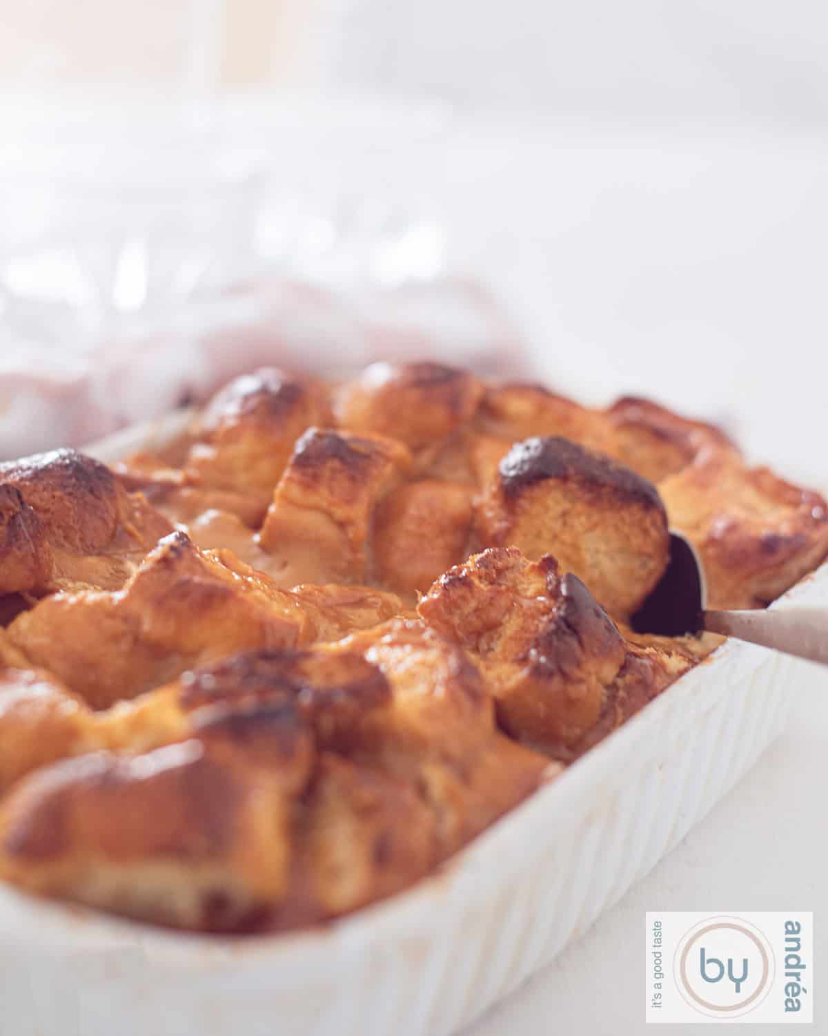 Een hoogtefoto van een witte ovenschaal gevuld met karamel broodpudding. Een lepel neemt een hap