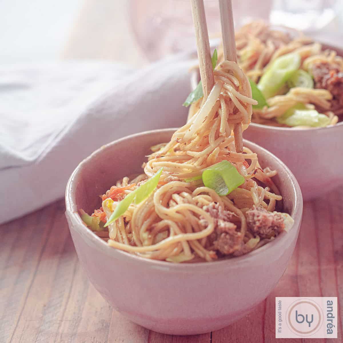 A square photo of two pink bowls filled with Chow Mein with ground beef and vegetables. Chopsticks grab noodles