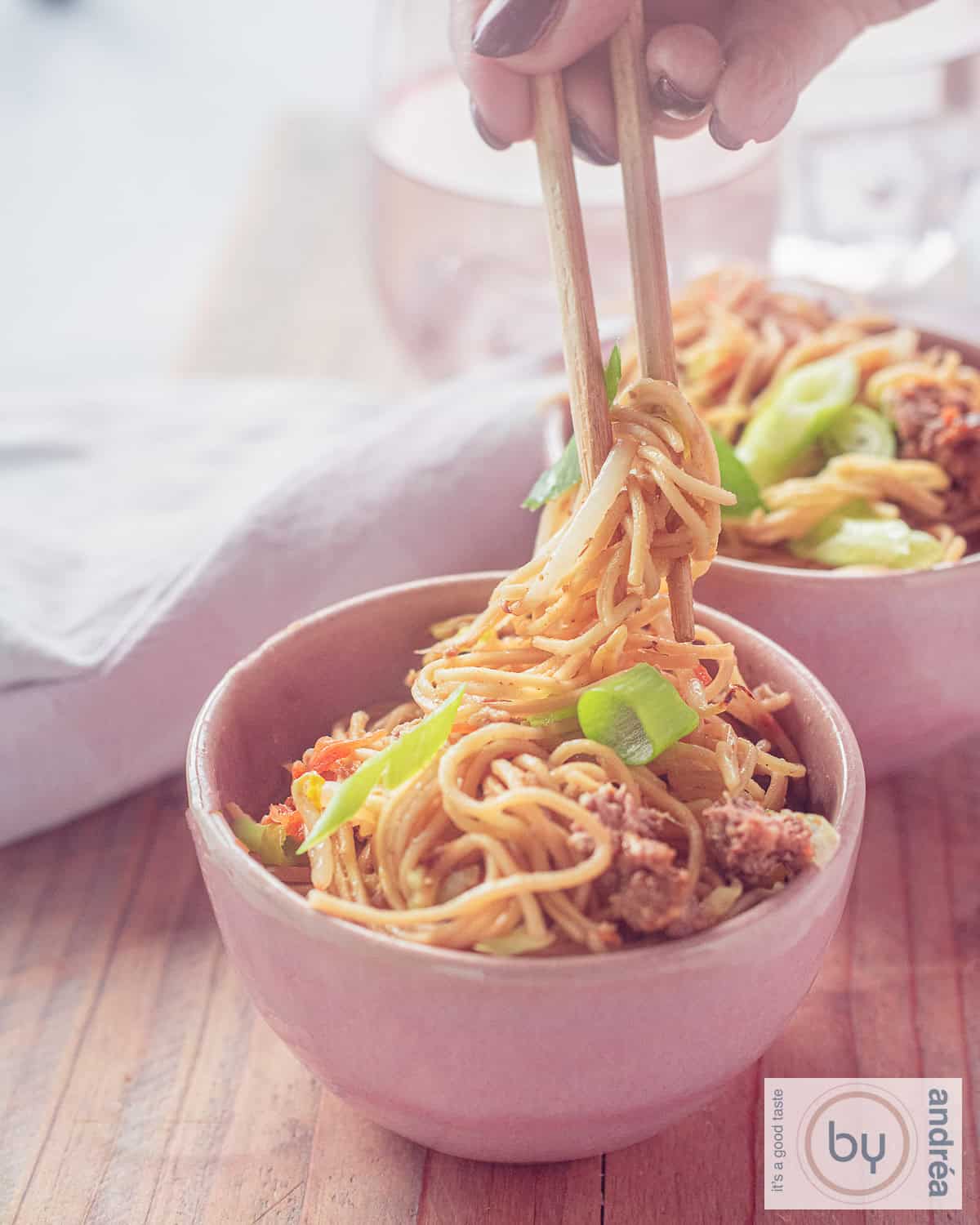Een hoogtefoto met twee roze kommen, gevuld met noedels, groenten en gehakt. Twee chopsticks pakken een hap noedels