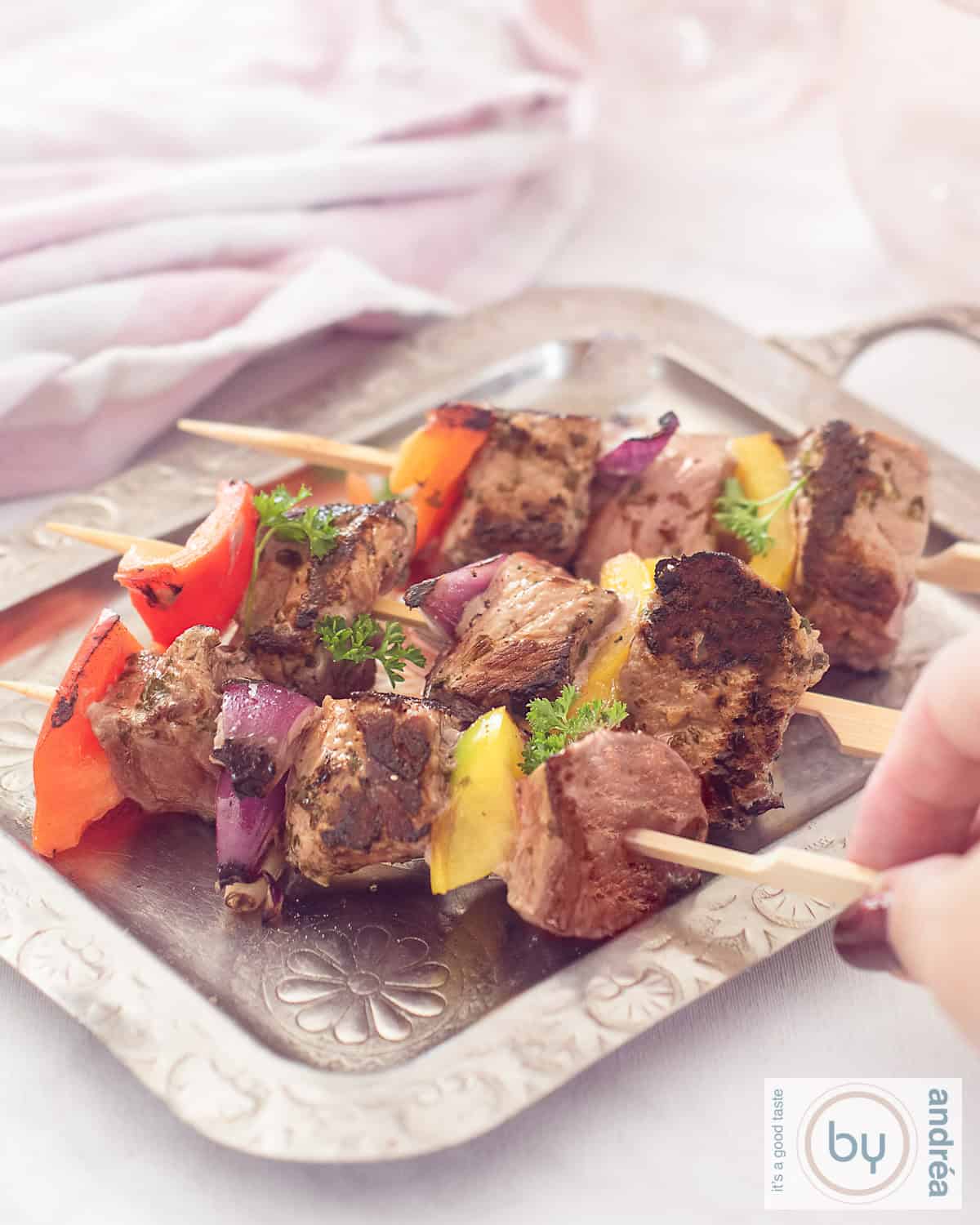 Een hoogtefoto met een zilveren schaal met drie spiesjes met biefstuk, ui en paprika. Een hand pakt een stokje.