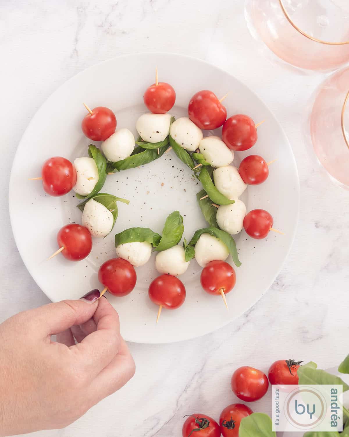 Een foto van bovenaf van een wit bord met ca. 12 spiesjes met tomaat, mozzarella en basilicum. Een hand pakt een stokje