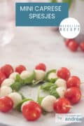 Een deel van een bord met spiesjes in Caprese stijl met tomaat, mozzarella en basilicum. Een tekst bovenin makkelijk recept, mini caprese spiesjes