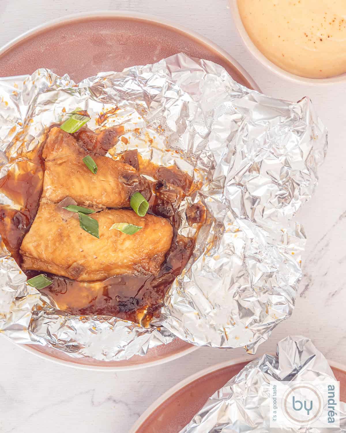 Een foto van bovenaf van een aluminiumfolie met pangsiusfilet in ketjapmarinade. Een bord met aluminiumpakketje onderin.