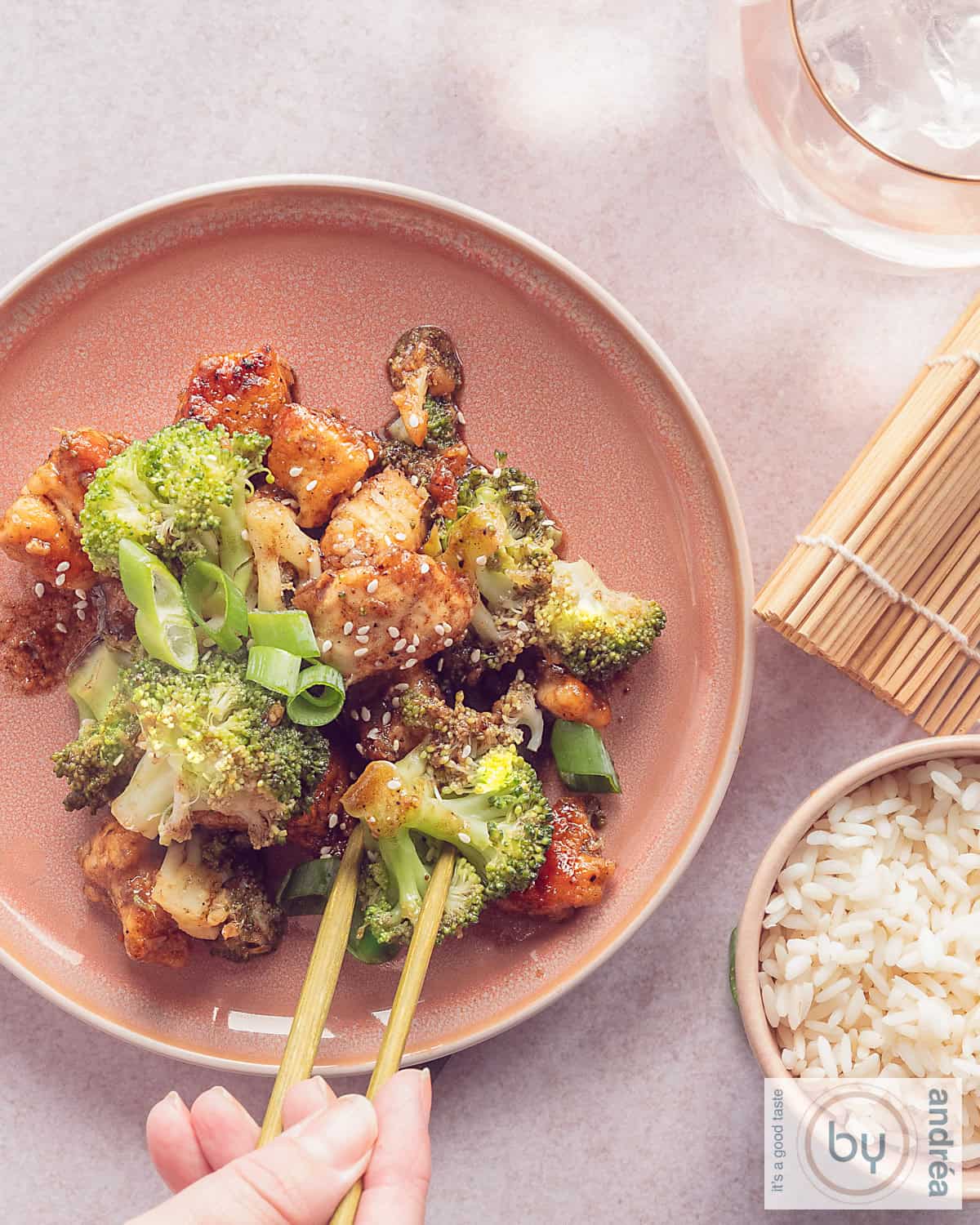 Een foto van bovenaf een roze bord met broccoli, kip in Aziatische saus geroerbakt. Twee chopsticks pakken een stukje. Een kom rijst linksonderin.