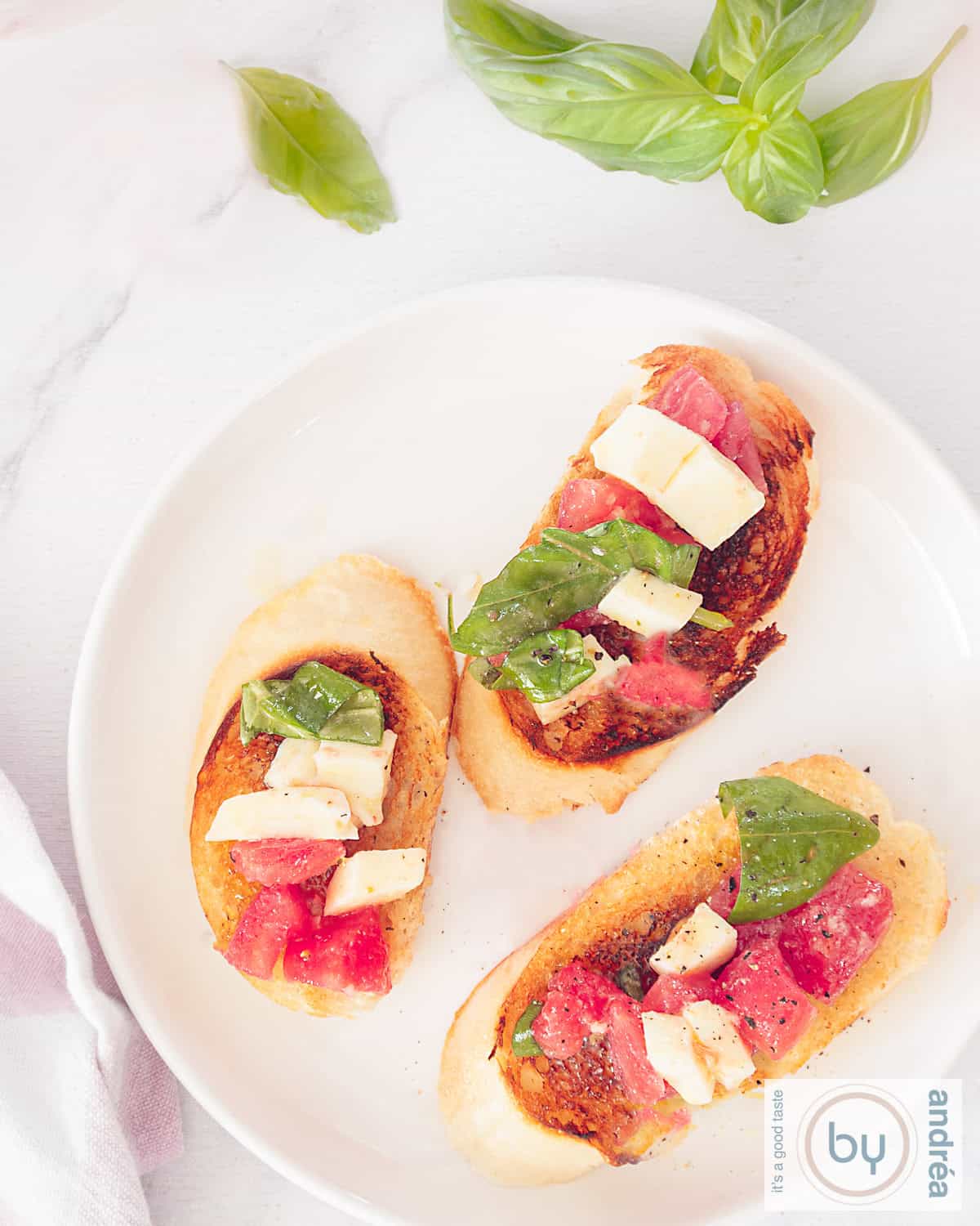 Een foto van bovenaf van een wit bord met drie stukjes gegrild stokbrood met een tomaten mozzarella salade. In de bovengrond blaadjes basilicum.