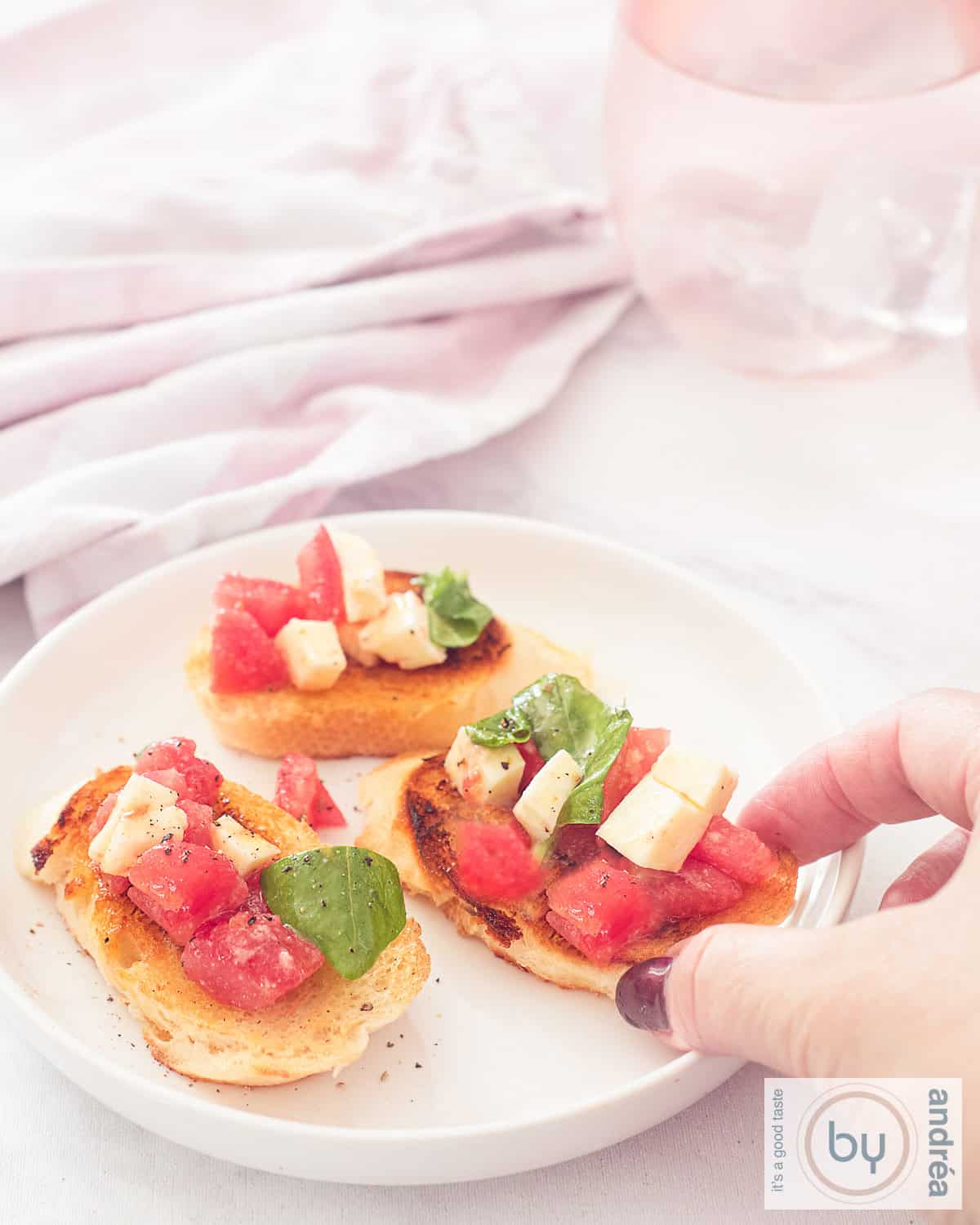 Een hoogtefoto met een wit bord met drie bruschetta stokbroodjes met tomaat, mozzarella en basilicum topping. Een hand pakt een bruschetta.