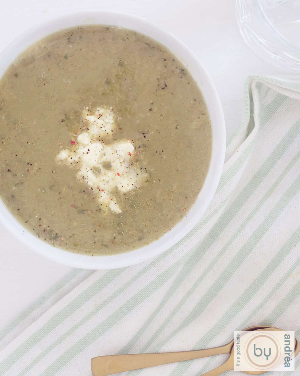 Een foto van bovenaf. Links bovenin een witte kom gevuld met romige courgettesoep met feta. Daaronder een groen wit gestreepte theedoek.