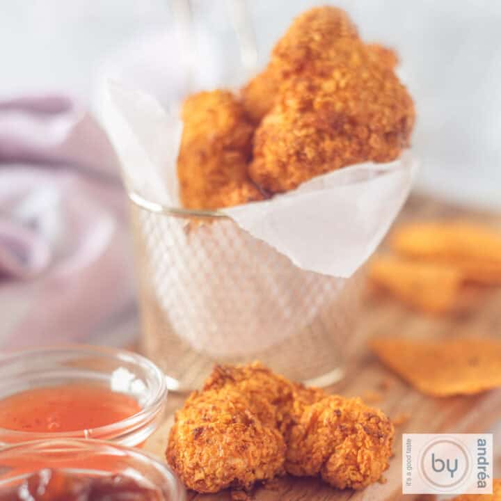 Een vierkante foto van een houten ondergrond met een bakje en extra losliggende kipnuggets met Dorito en sausjes.