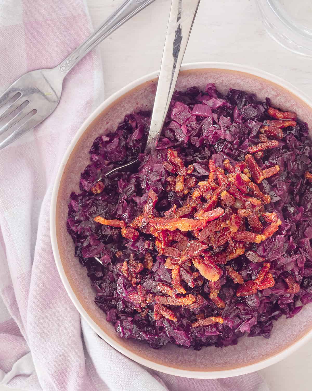 A photo from above of a pink bowl with red cabbage and a topping of bacon. A white background and on the left a pink and white tea towel.