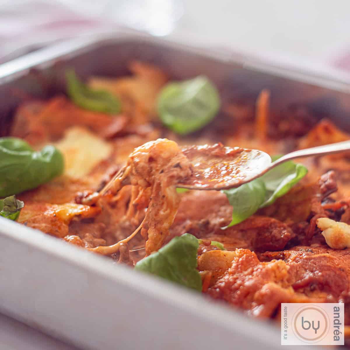 Een vierkante foto van een braadslede gevuld met cheesy eenpans lasagne met mascarpone en gehakt. Een lepel neemt een hap.