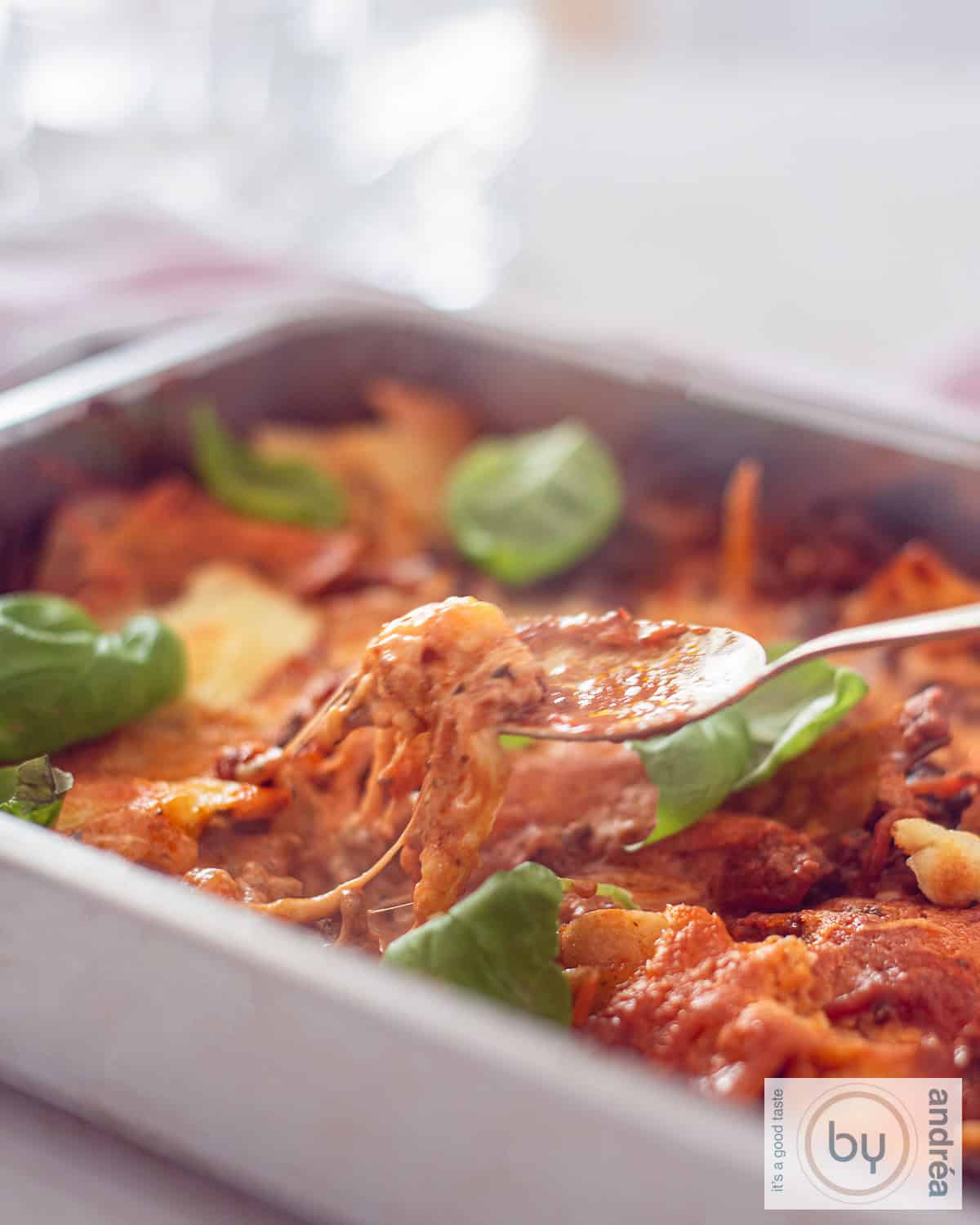 Een hoogtefoto met een braadslede gevuld met eenpans lasagne met mozzarella, mascarpone, gehakt en tomatensaus. Een lepel neemt een hap.
