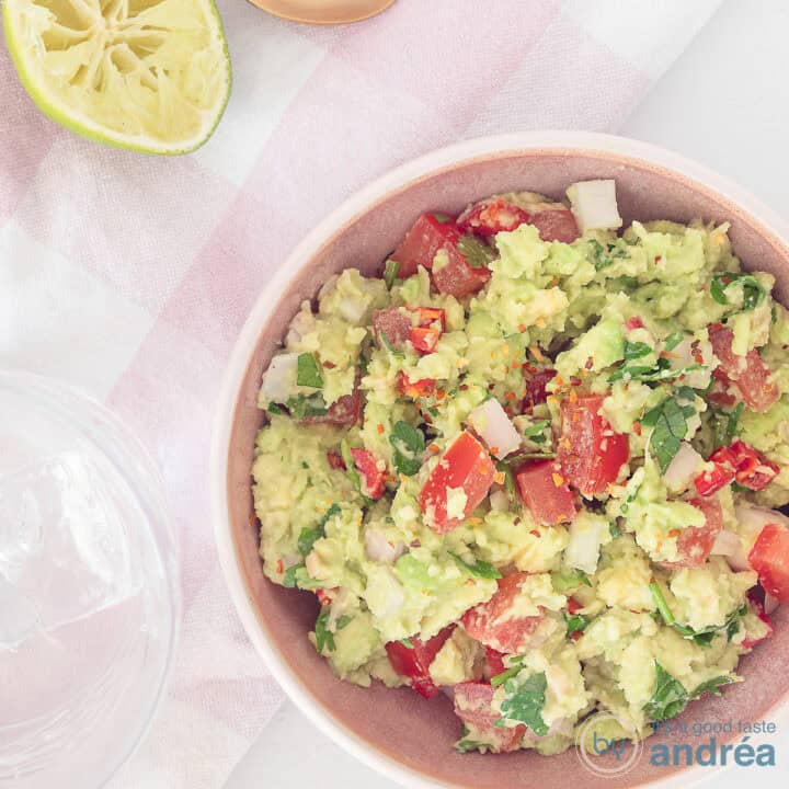 Een vierkante foto van bovenaf genomen. Rechts onderin een schaal met zelfgemaakte guacamole met avocado, tomaat en limoen. Bovenin is nog een stukje limoen zichtbaar.