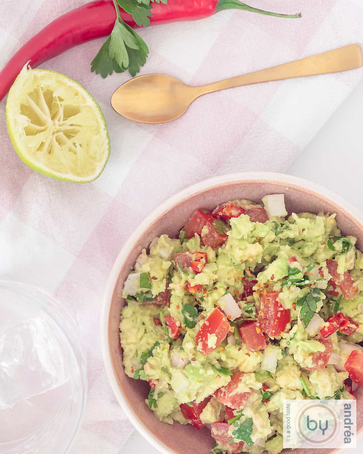 Een foto van bovenaf, rechts onderin een kom gevuld met guacamole. Bovenin een roze witte theedoek met daarop een gouden lepel, een uitgeknepen limoen, peper en koriander.