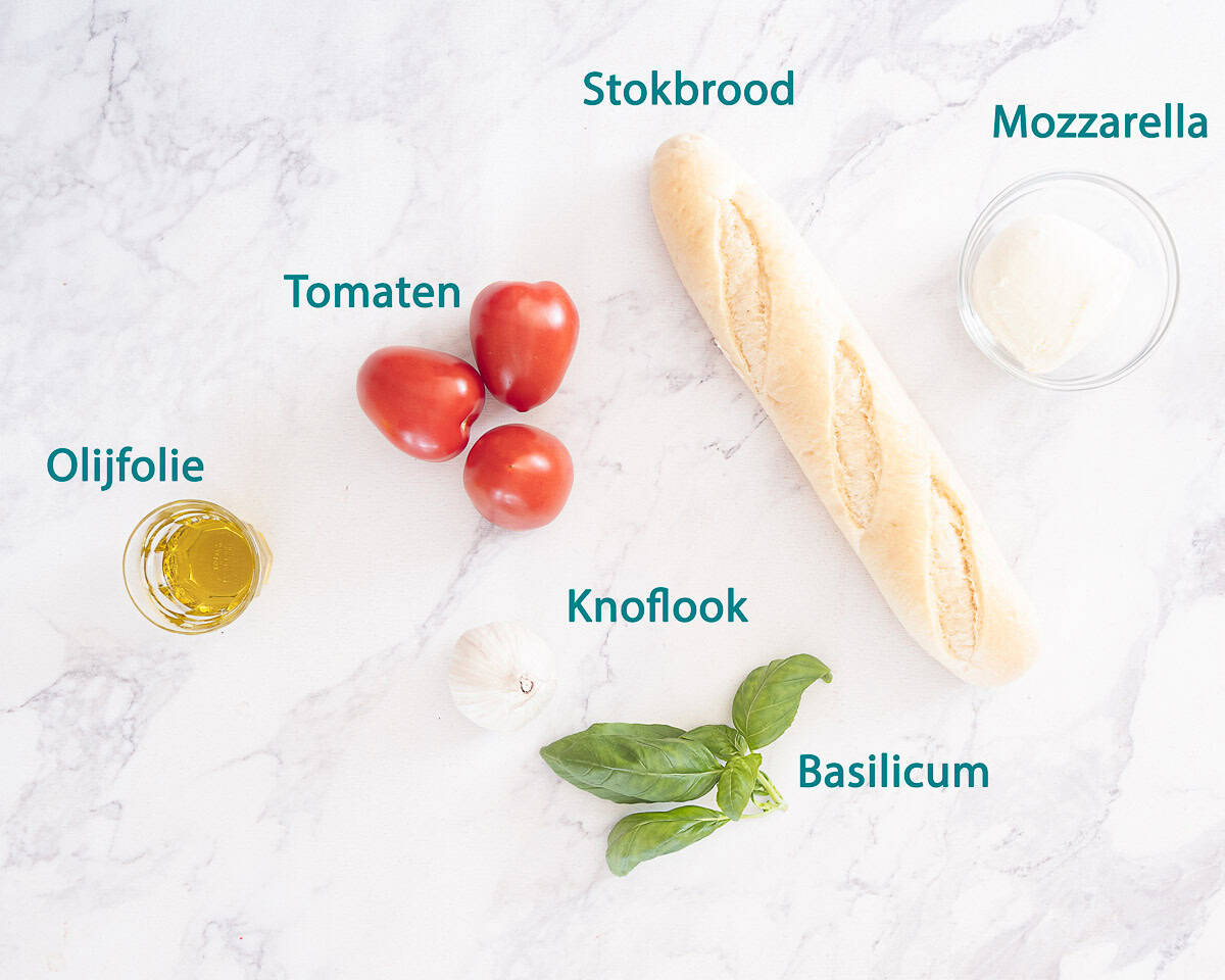 Ingredienten Bruschetta met Tomaat, Mozzarella en Verse Basilicum op een gemarmerde witte ondergrond.