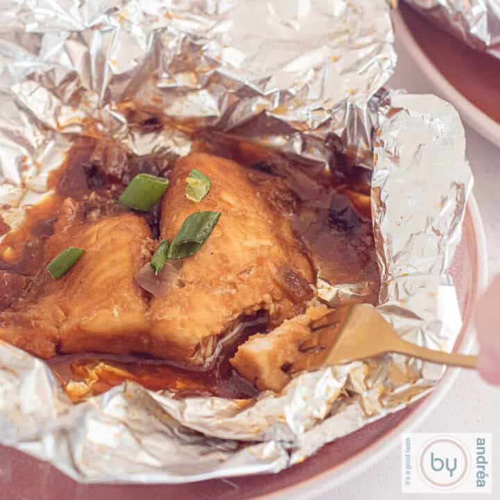 Een vierkante foto van een roze bord met opengevouwen aluminiumfolie met daarin pangasiusfilet van de BBQ. Een vork prikt een stukje eraf.