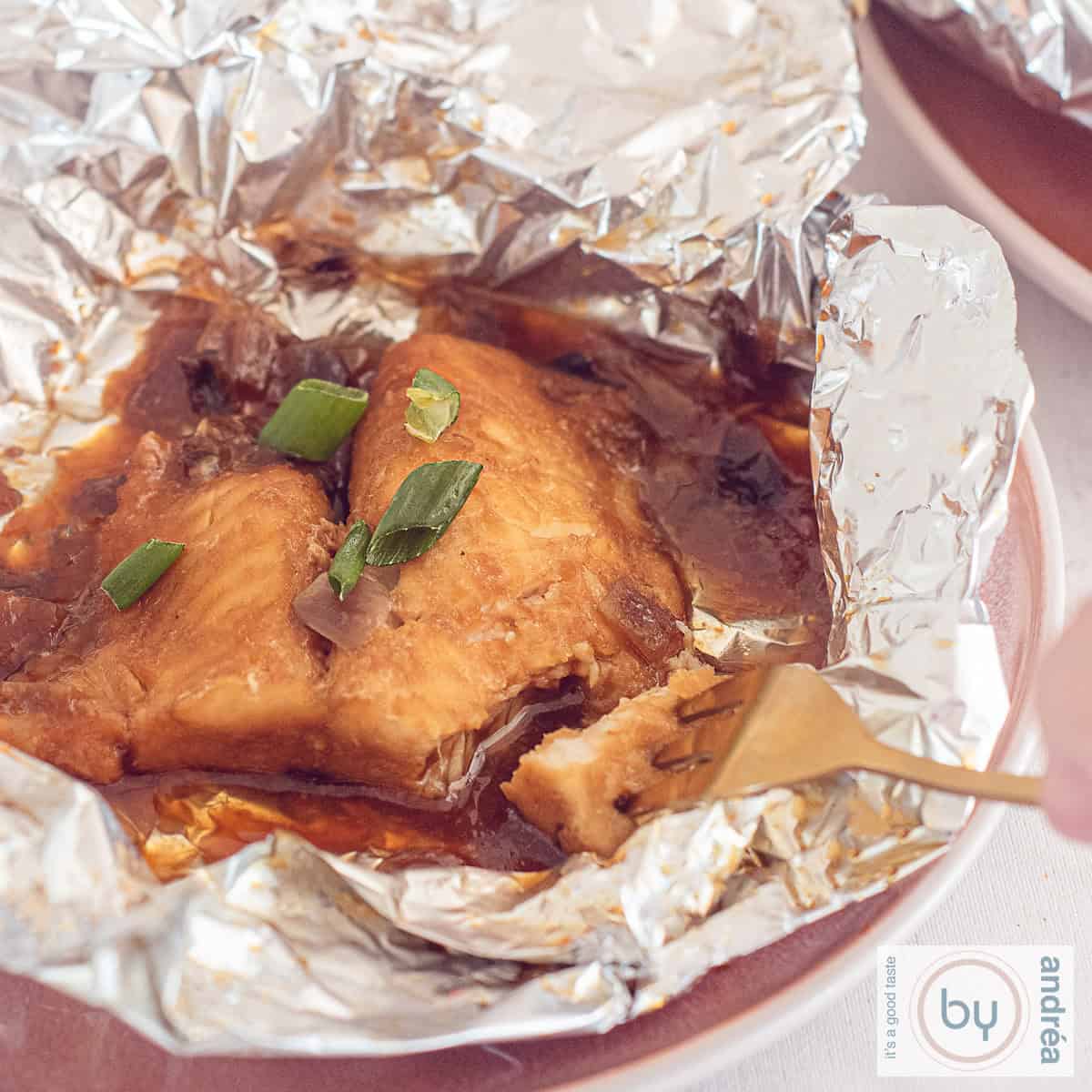 Een vierkante foto van een roze bord met opengevouwen aluminiumfolie met daarin pangasiusfilet van de BBQ. Een vork prikt een stukje eraf.