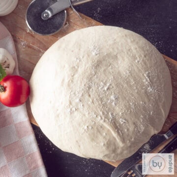 Een vierkante foto van bovenaf genomen van een bol pizzadeeg op een houten plank. Een tomaat en knoflook en een pizzasnijder liggen erbij.